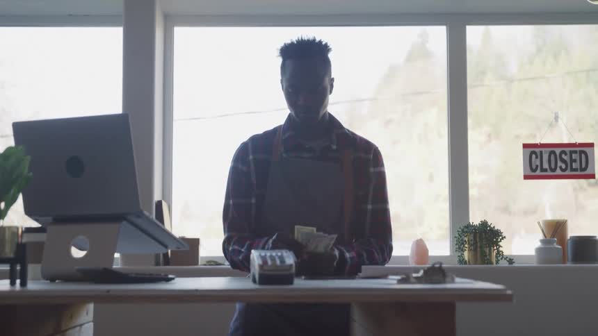 A man counting cash at a table with a laptop and a credit card machine