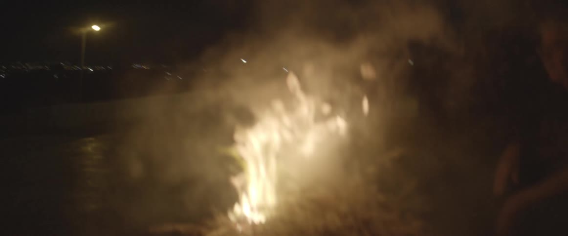 A man is squatting next turn a burning pile of twigs at night