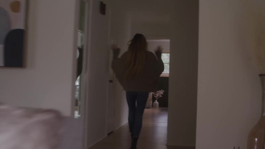 A woman with long blonde hair wearing a beige cardigan and jeans walks from a bright living room down a hallway and closes a door behind her