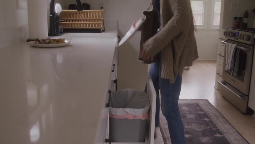 A woman in the kitchen tosses a meal from a plate into the bin then puts the plate into the sink at midday