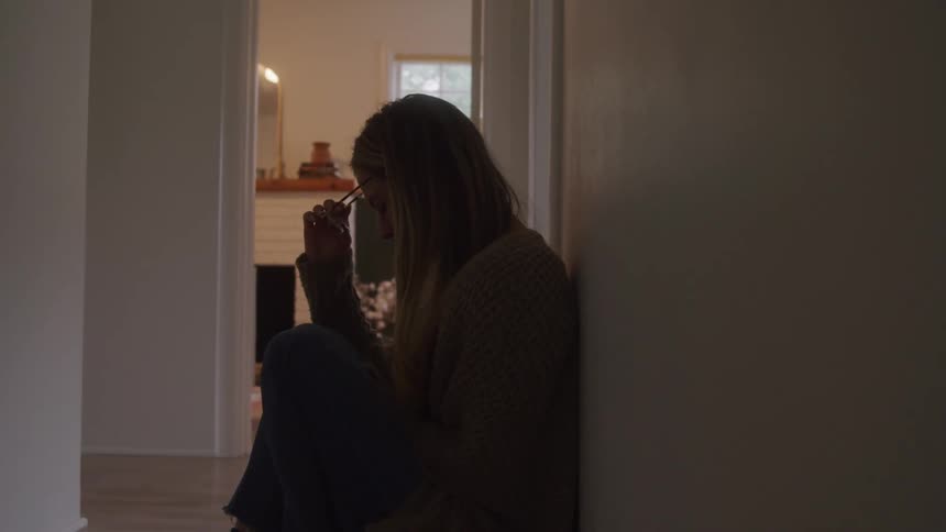 A woman leaning against the wall sits on the floor and breaks down in sadness