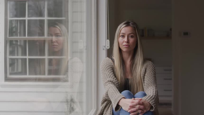 A young woman with blonde hair sits indoors beside a glass window wearing a brown sweatshirt