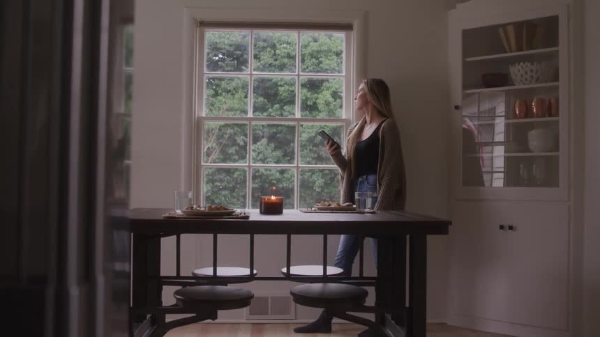 A woman standing by a window holding a phone looking outside then looking at the phone