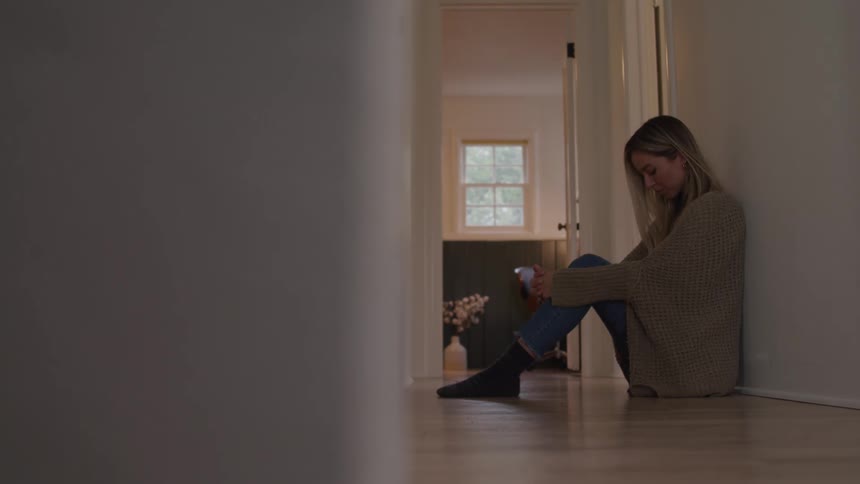 Woman sitting on the hallway floor leans against the wall holding a mug with a thoughtful gaze