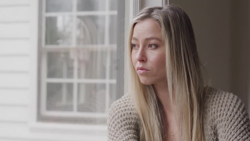 A young woman with long blonde hair sits beside a glass window at midday looking sad and wearing a brown knitwear shirt