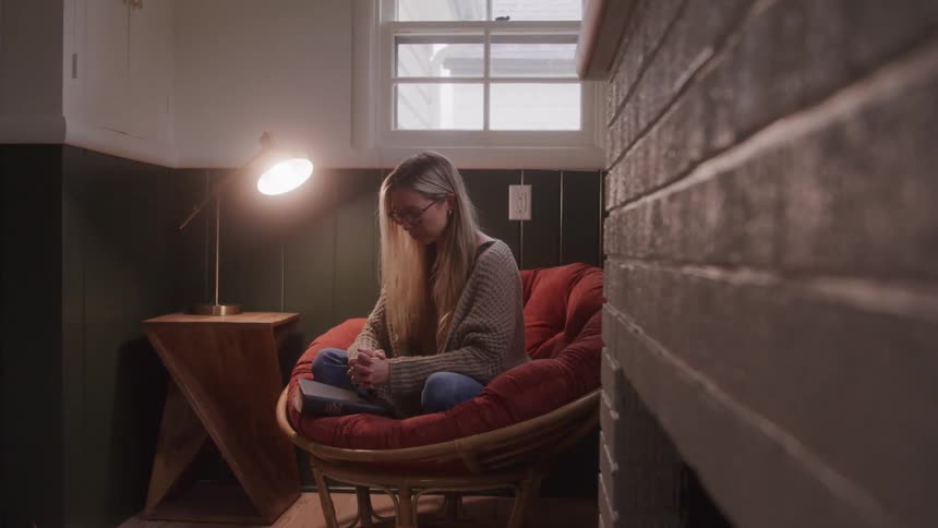 A young adult woman with long blond hair and glasses sits in an orange chair looking down at a book in her lap with a lit lamp on a small wooden table to her left and a window in the background