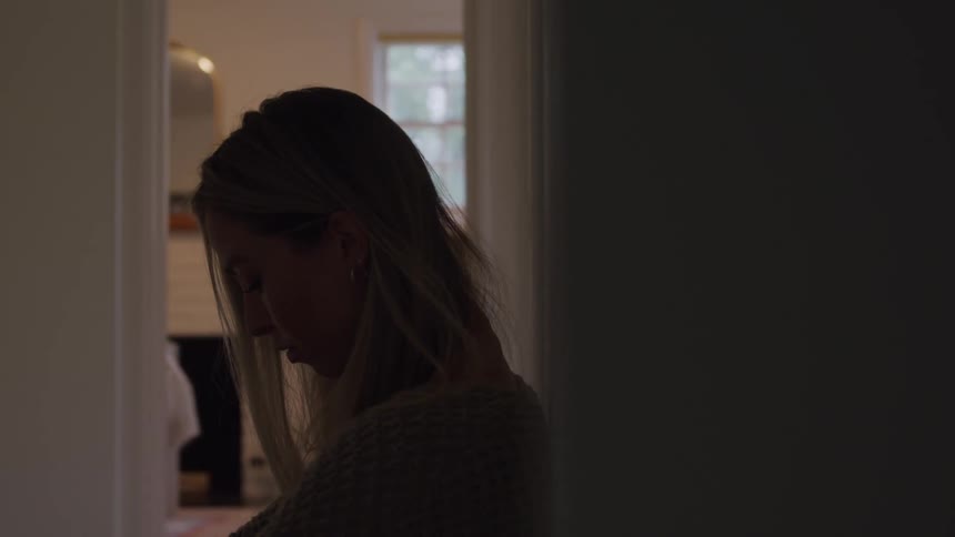 A young woman with long hair leans against a wall indoors looking sad