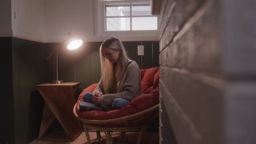 A young adult woman with long blond hair and glasses sits in an orange chair looking down at a book in her lap with a lit lamp on a small wooden table to her left and a window in the background