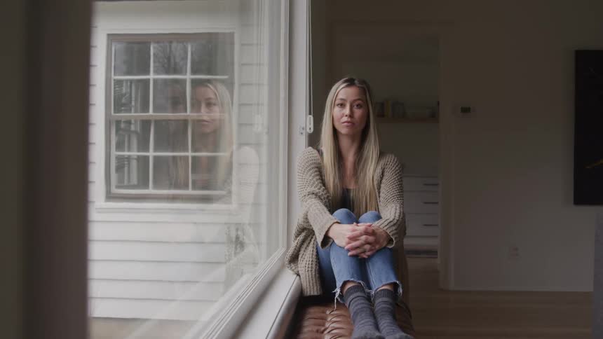 A woman sits at a picture window and looks ahead in thought