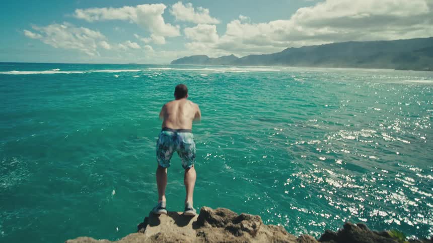 A man wearing trunks jumps from a rocky cliff into the ocean on a sunny day
