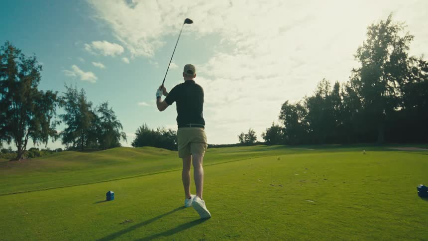 A man in a hat and golf attire swings a golf club on a green course under a bright cloudy sky then watches the ball fly before bending down to pick up his tee