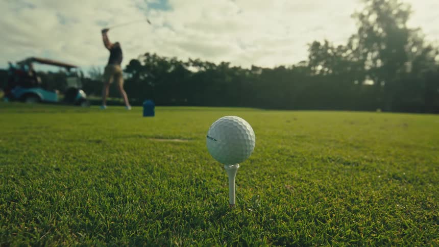 A hand wearing a blue wristband placing a golf ball on a tee in lush green grass with a blurred golf cart and another person in the sunny background