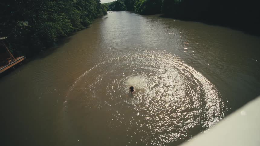 Person jumps off a bridge over a river surrounded by vegetation