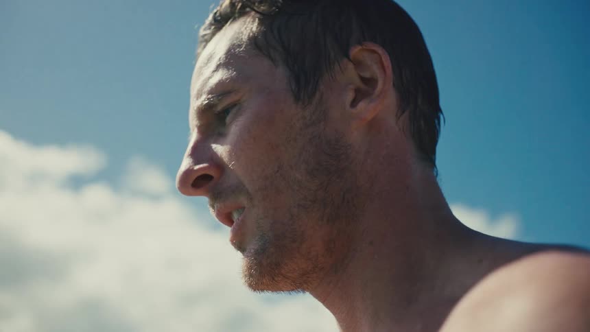 A close view of a mans face seen from the side with a slight stubble looking around against a bright blue sky with white clouds