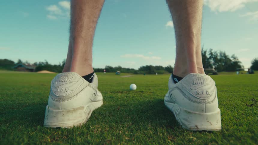 A lowangle shot from behind a persons legs showing their white nike golf shoes as they address a golf ball with a club on a green course under a partly cloudy sky