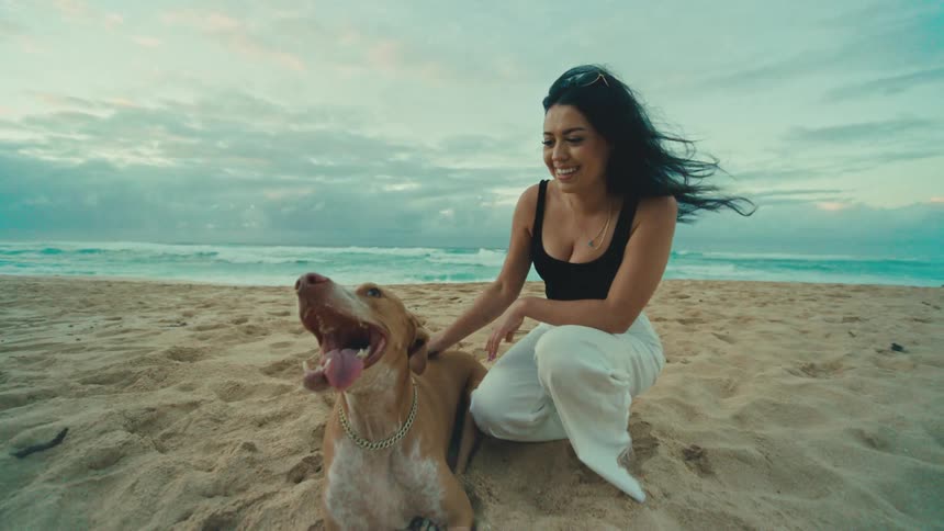 The woman is petting her dog in the sand