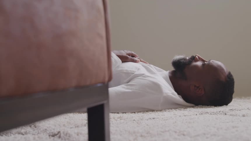 Man lying on his back on the carpet keeps his hands on his chest next to an upholstered piece of furniture