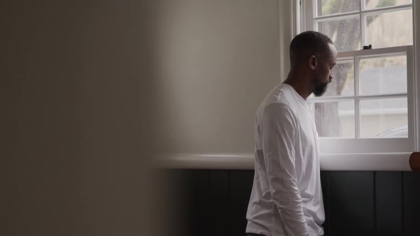 An adult man with a beard wearing a white long sleeved shirt looking out of a window with a somber expression