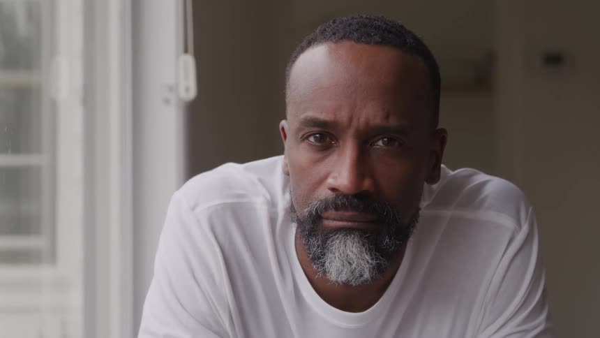 A man with a white and black beard stands indoors beside a window at midday wearing a white tshirt