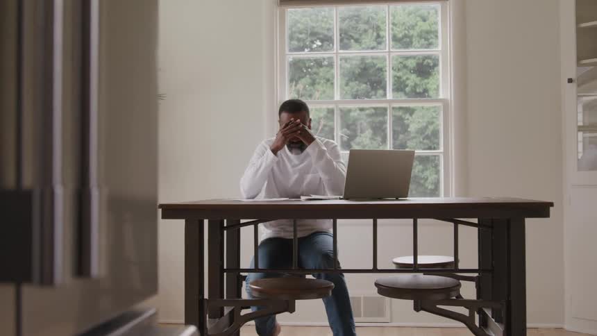 A middleaged man sits behind a desk at midday looking sad with a computer in front of him and both hands on his forehead elbows resting on the table