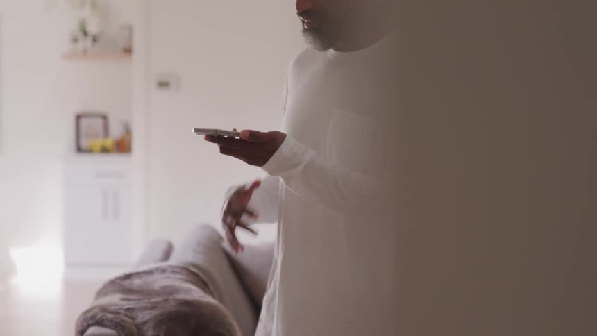 An adult man with a beard wearing a white long sleeved shirt and dark pants stands holding a phone and gesturing with his free hand while looking at the device in a home setting