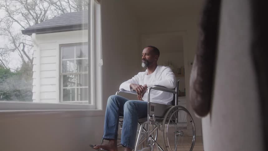 A man sits in a wheelchair in front of a large picture window in a house