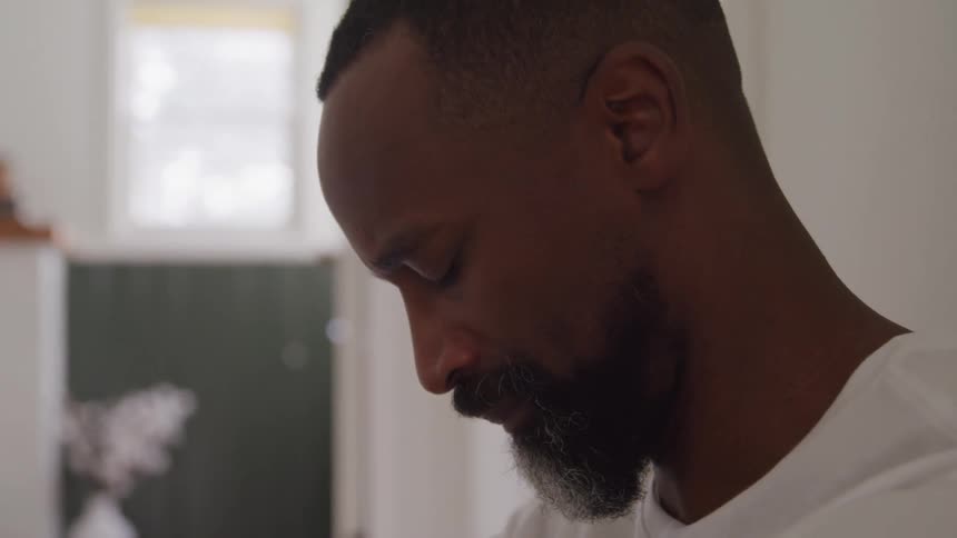 A sad man sits against a wall in a house and slowly lowers his head