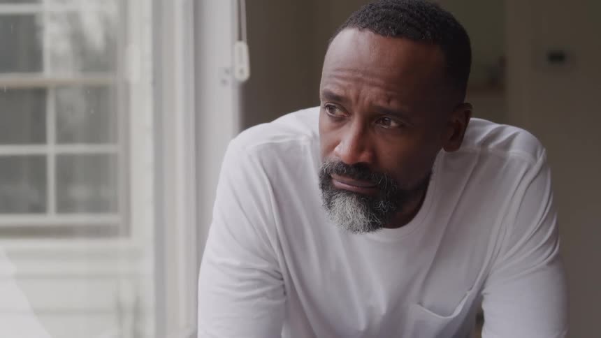 Graybearded man in a white shirt gazes thoughtfully out the window