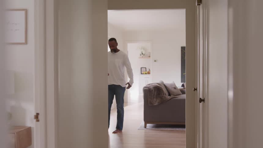A man wearing a white longsleeve tshirt and jeans walks back and forth inside his living room at midday angrily talking on his phone