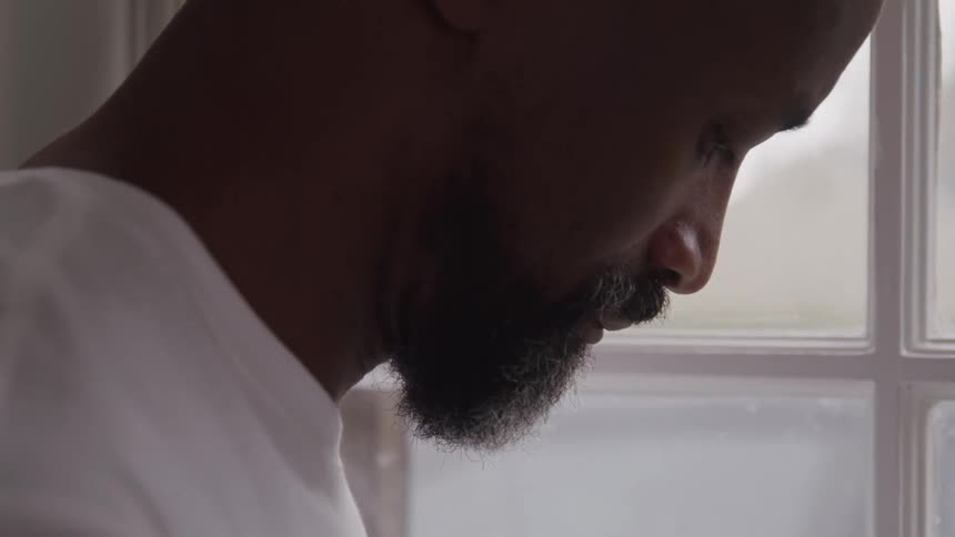 An adult man with a beard looks down with a somber expression seen in profile while standing in front of a window with white frames wearing a light colored shirt