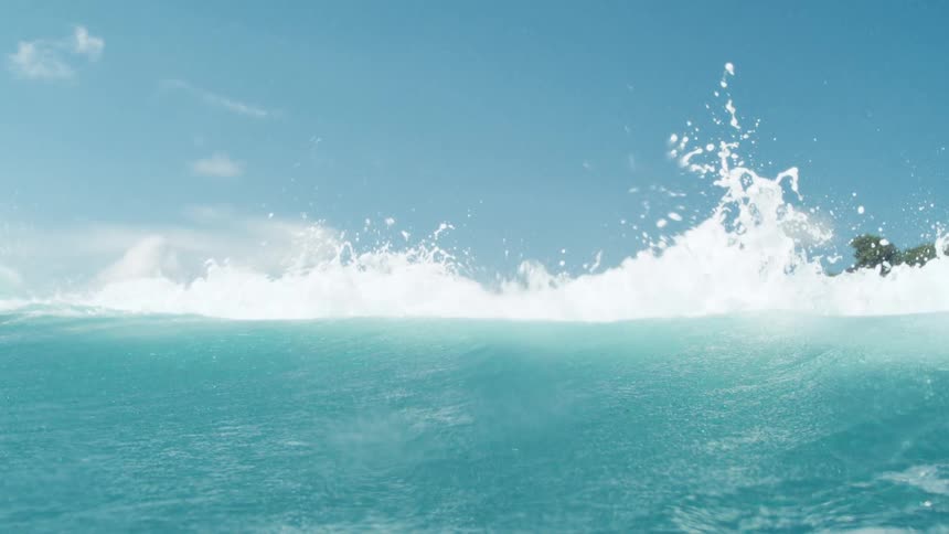 A turquoise ocean wave cresting and sending white spray into the clear blue sky with a distant land on the right