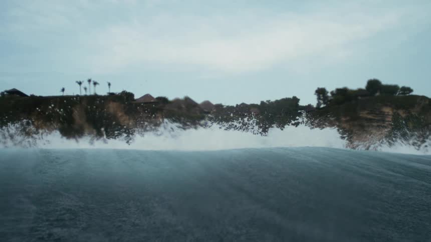 Wave breaks near the shore with cliffs and palm trees in the background under a cloudy sky