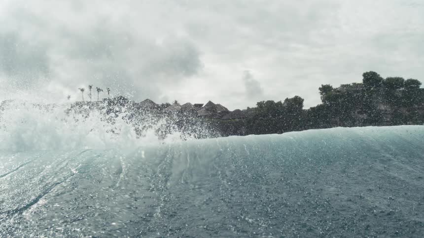 Wave rises in front of a hillside with vegetation and houses in the background on a cloudy day