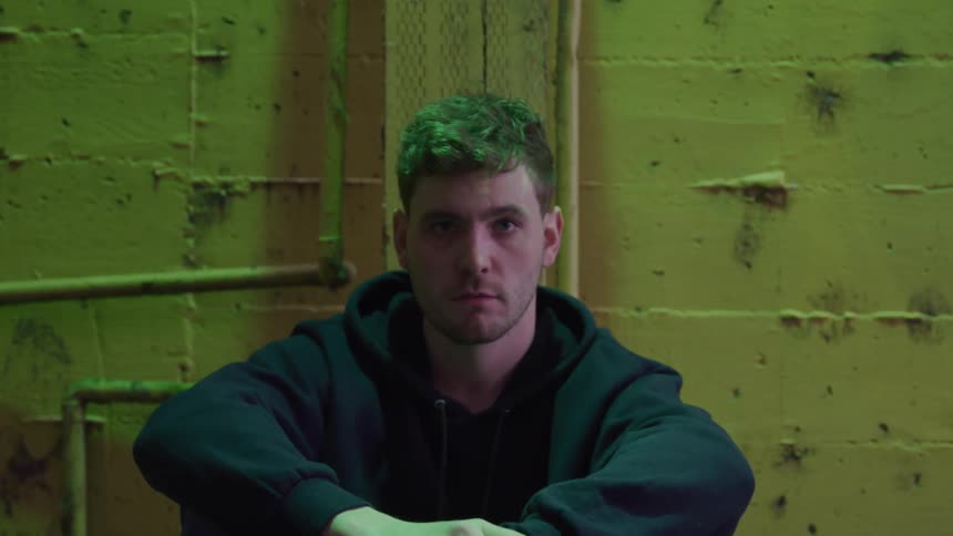 Man in a dark hoodie sits with arms resting on his knees in front of a yellow wall with pipes