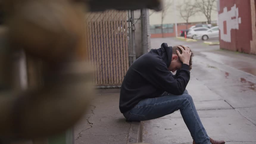 A young adult man sits on a curb with his head in his hands appearing distressed in a street with parked cars and a red building visible in the background