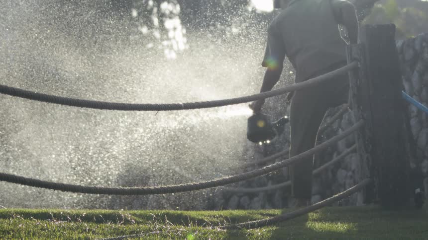 A worker sprays water onto grass using a pressure washer creating a mist in the sunlight
