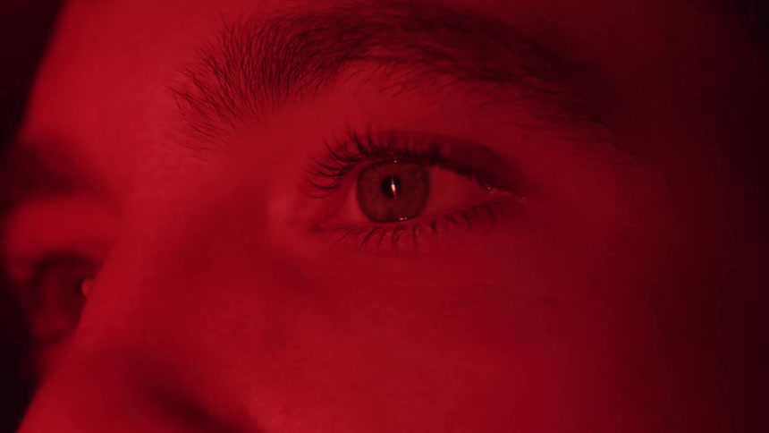A closeup of a persons eyes are lit by red light as they blink and gaze off to the side