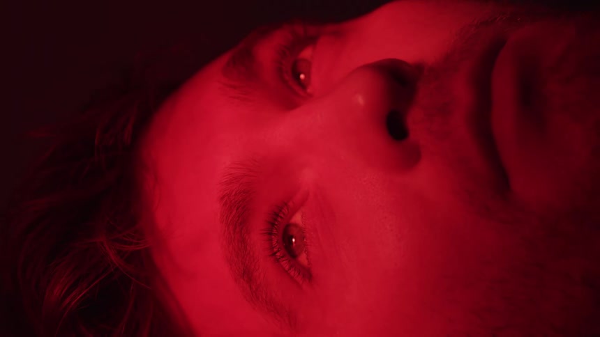 A close view shows a young adult persons face bathed in red light with their eyes opening revealing a pensive expression