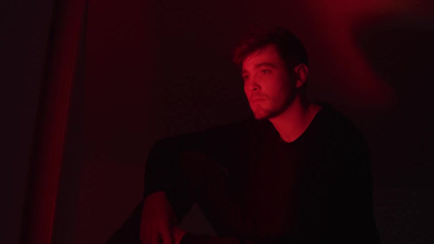 A young man sits in a dark room illuminated by a strong red light from the left with his head leaning on one hand and a somber expression on his face