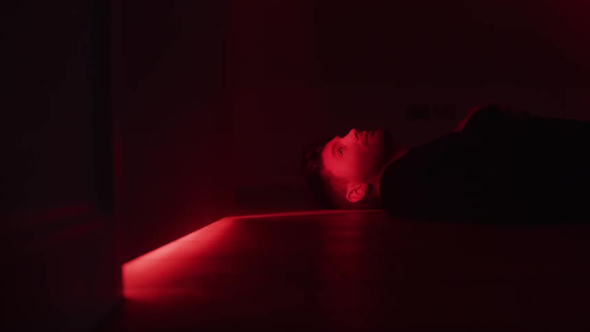 A man lies still on a dark surface illuminated by a strong red light emanating from under a door to the left casting a bright red glow across the surface and part of his face