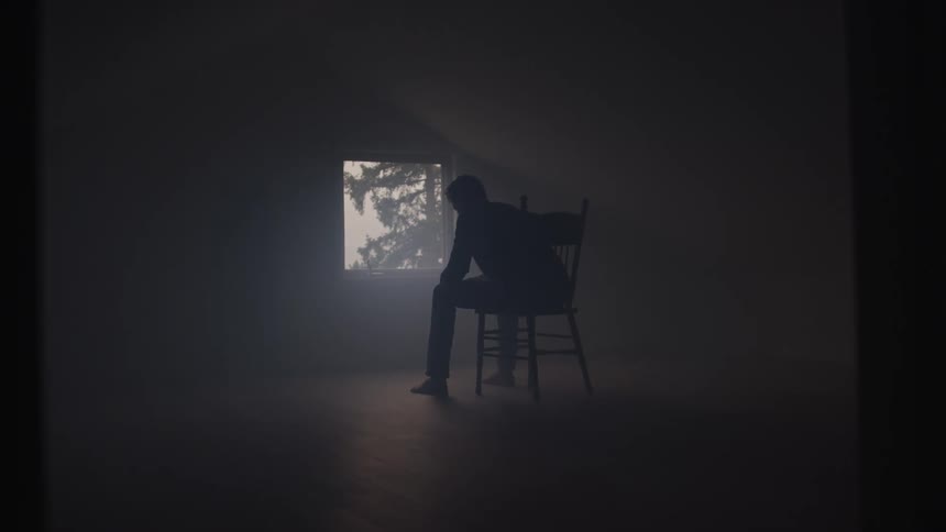 A silhouette of a person sits on a chair in a dark room facing a bright window that looks out onto a hazy outdoor scene