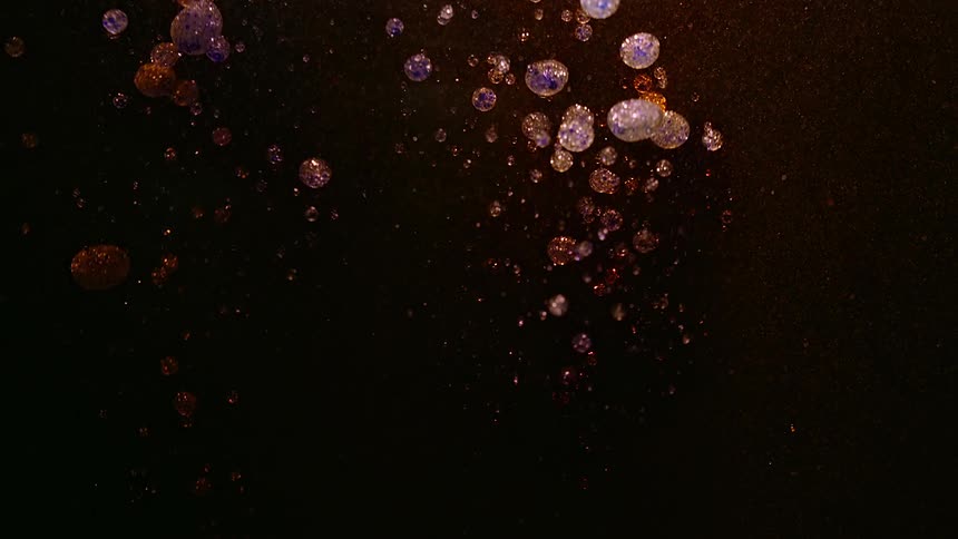 Iridescent oil bubbles float against a dark backdrop after something drops into the liquid illuminated with golden and purple light
