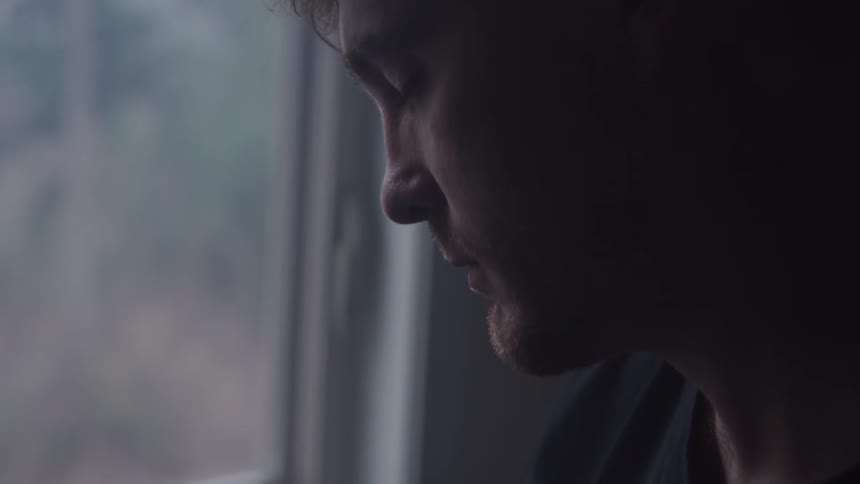 Man with an introspective expression stands silently by the window lit by natural light