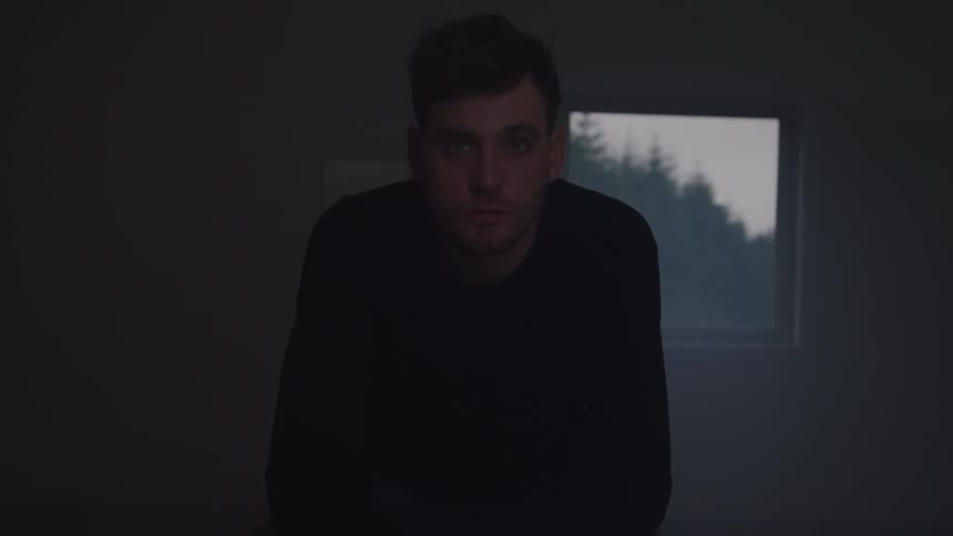 A young adult man sits in a dark room looking directly at the camera with a solemn expression with a window showing dark trees outside in the background