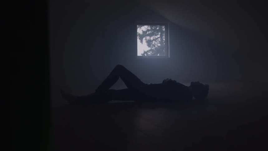 A woman lies on the floor in a dark empty room at sunset with light streaming in through a small window