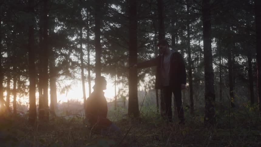 A man kneels in the woods at sunset with his eyes blindfolded while another man stands nearby holding a gun to his head surrounded by trees