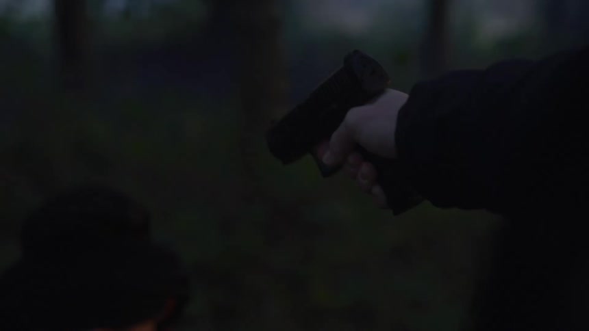 A man points a gun at the head of a fearful teenage boy kneeling on the ground in the woods