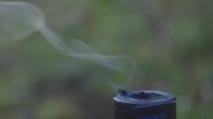 Smoke rises from a small tubular burning object outside in the grass