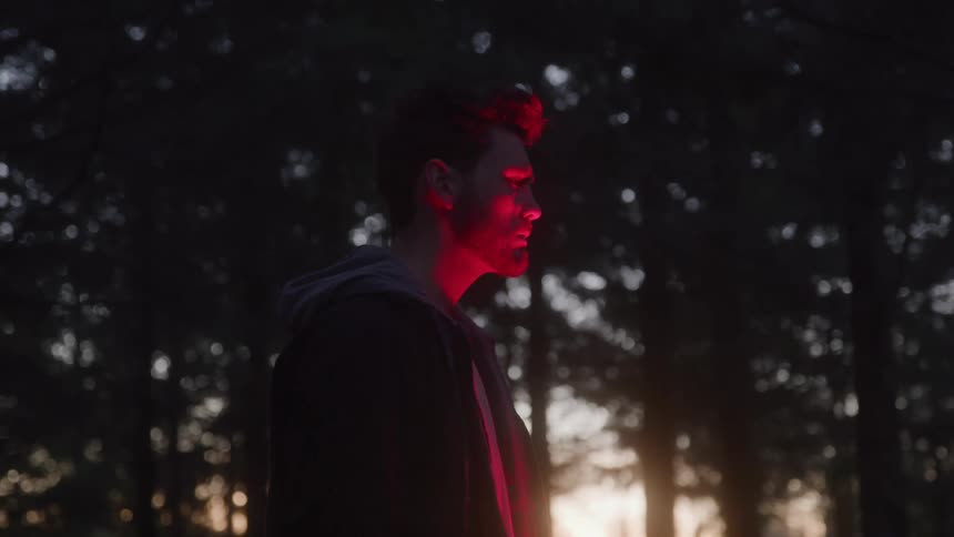 A man wearing a jacket stands beside a campfire in the woods at sunset