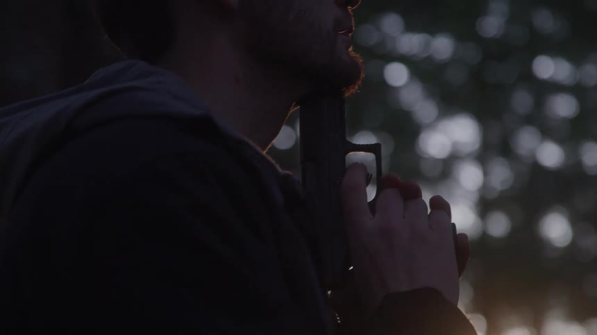 A man with a beard silhouetted against a bright hazy background of trees holding a handgun with both hands suggesting a focused or tense moment